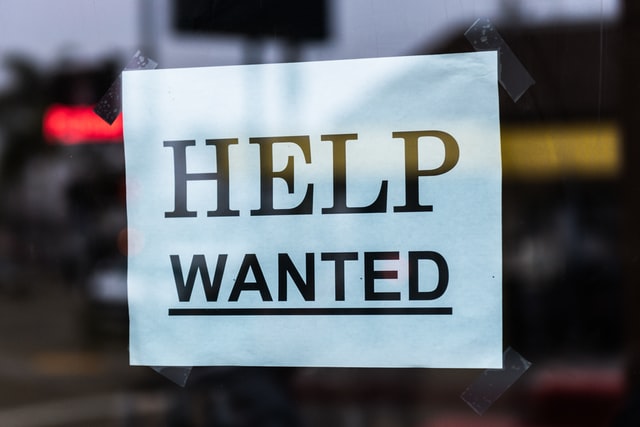 A sign hanging on a glass window that reads "Help Wanted"