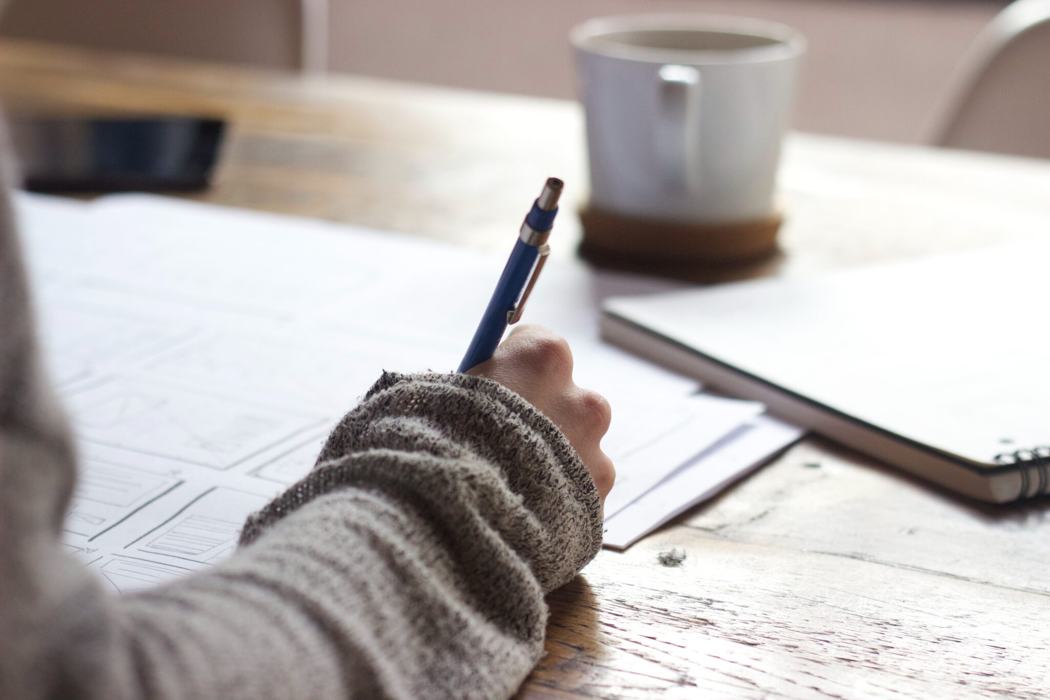 A person sitting a desk holding a pen. A coffee cup is in front of them as well as a notepad and laptop computer.